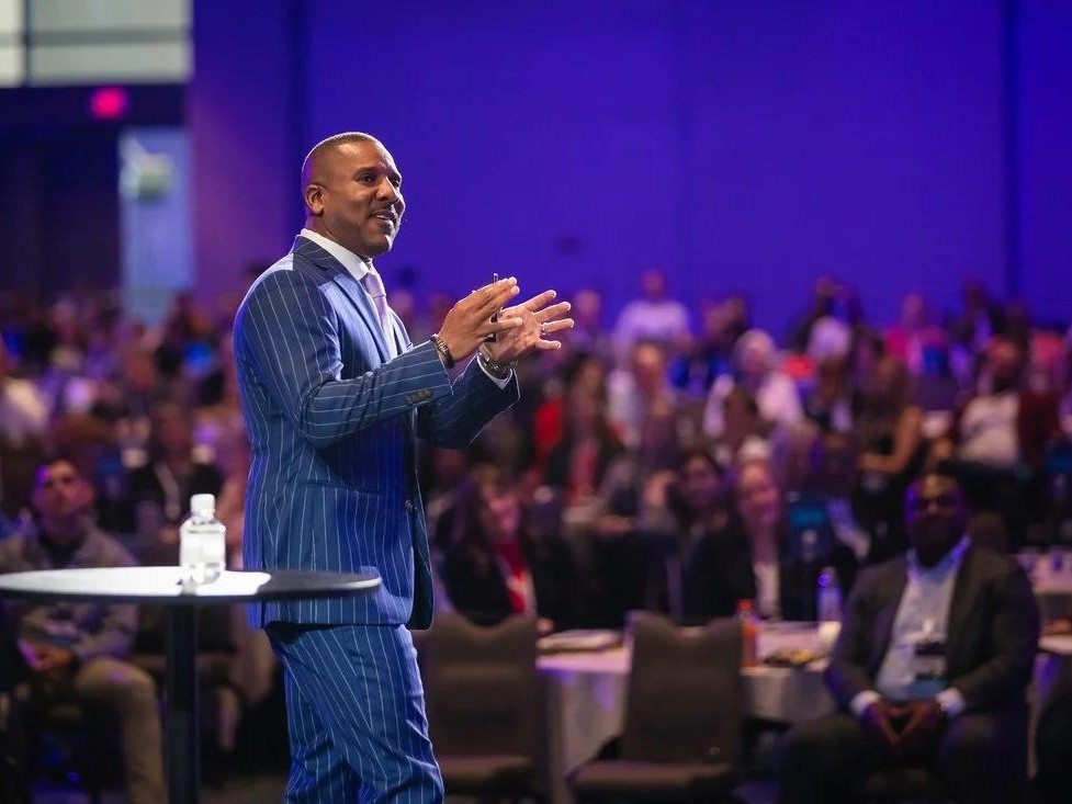 A keynote speaker addressing a large conference audience under stage lighting